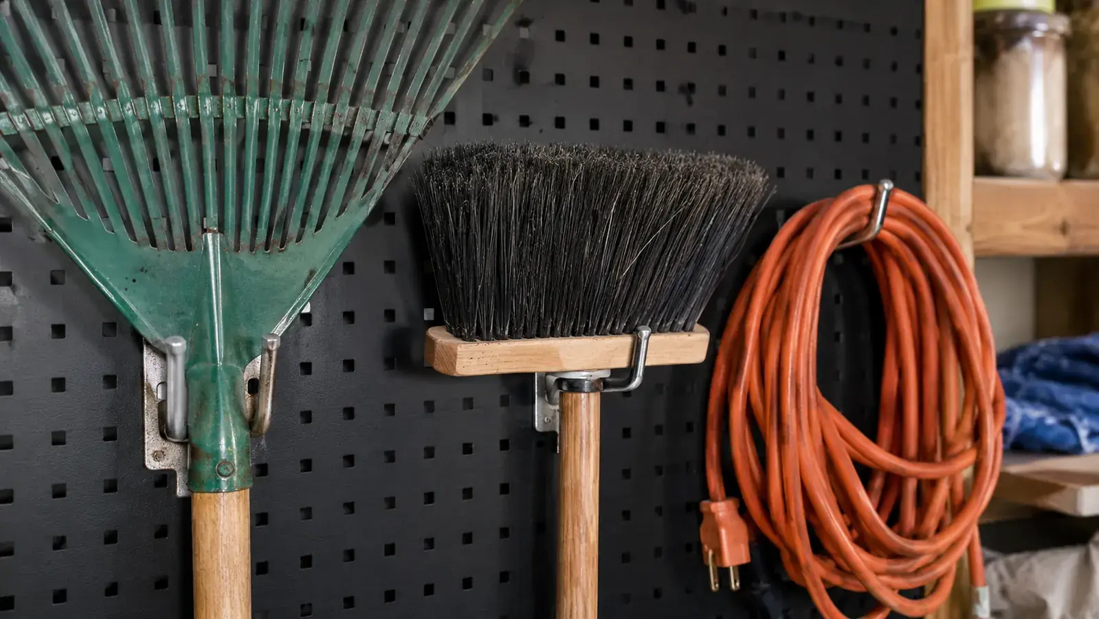 Close-up of metal pegboard hooks holding a rake, a broom, a ladder, and looped extension cords in a garage