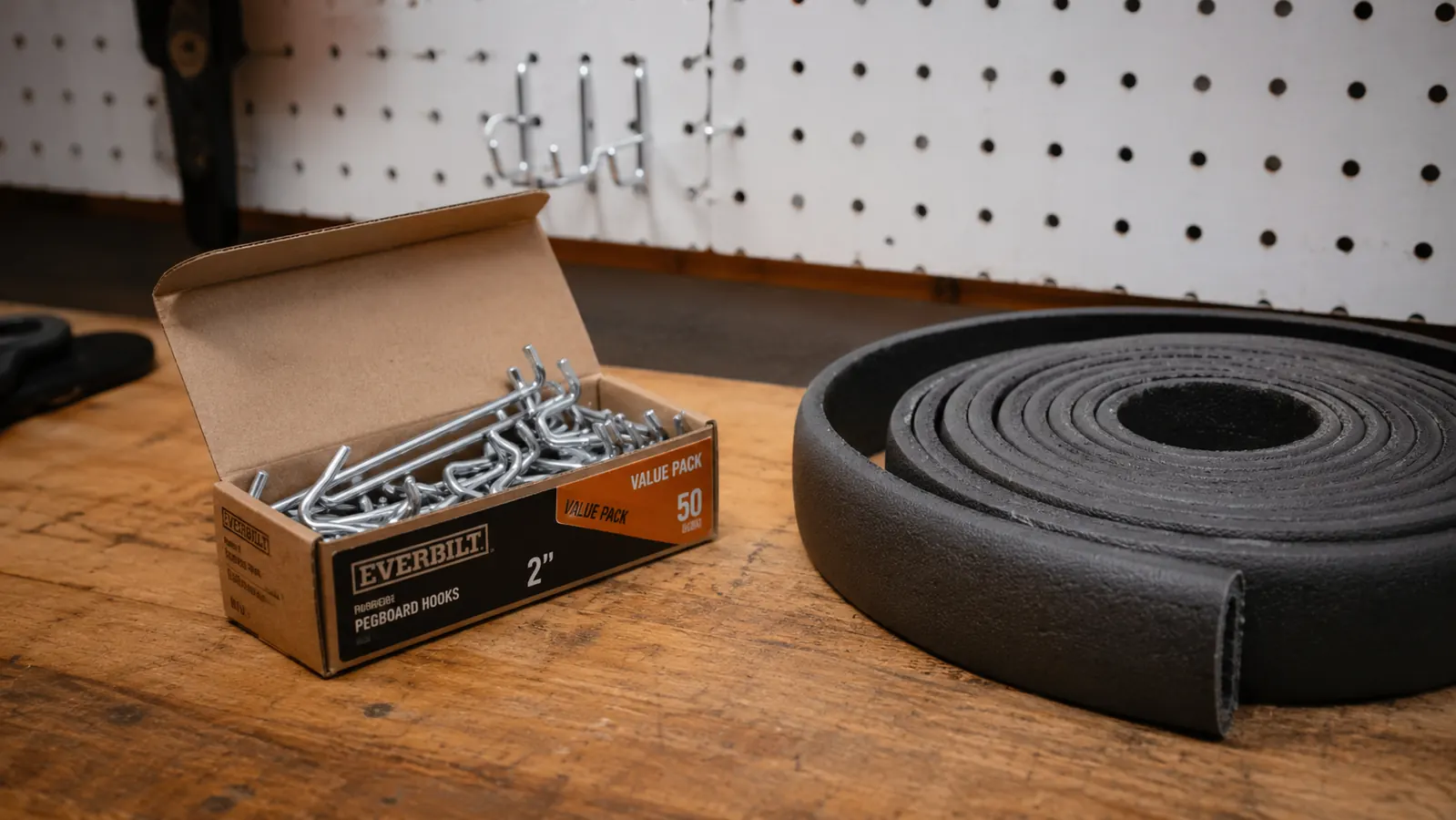 A small cardboard box of extra pegboard hooks and a roll of foam edge trim sitting on a workbench in front of the mounted pegboard wall