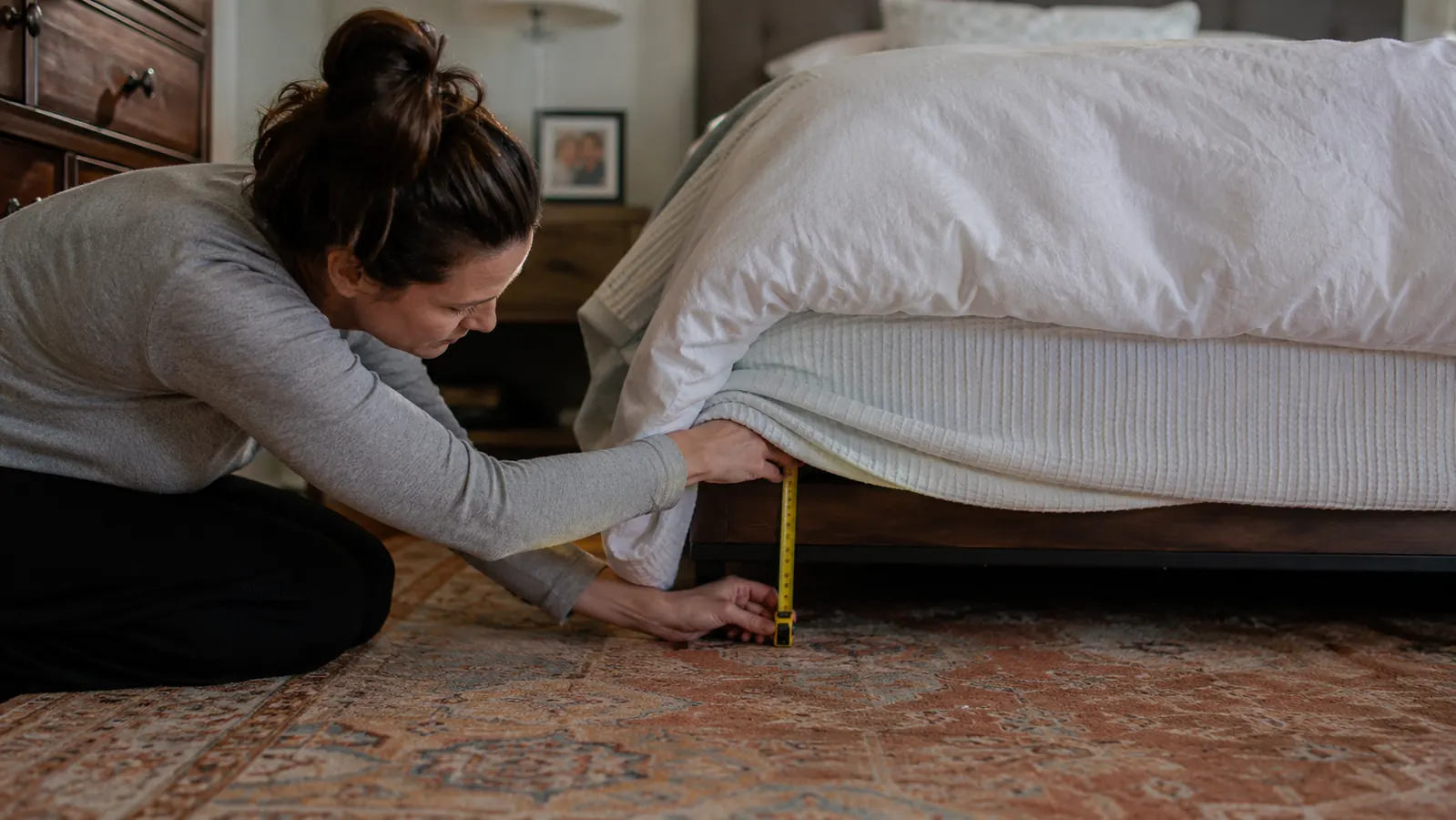A woman kneeling beside a bed checking the gap under a dust ruffle with a tape measure before sliding a storage bin underneath