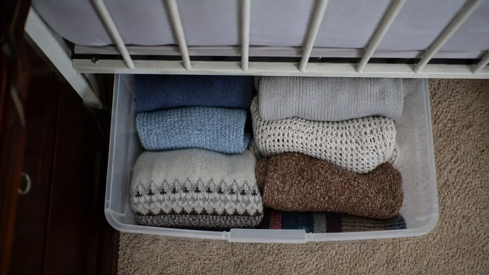 Overhead view of folded off-season clothing and blankets packed neatly inside a clear bin under a bed